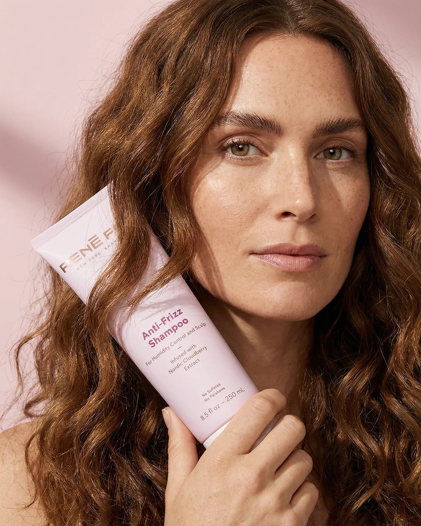 Woman holding a bottle of René Fris Anti-Frizz Shampoo against a pink background