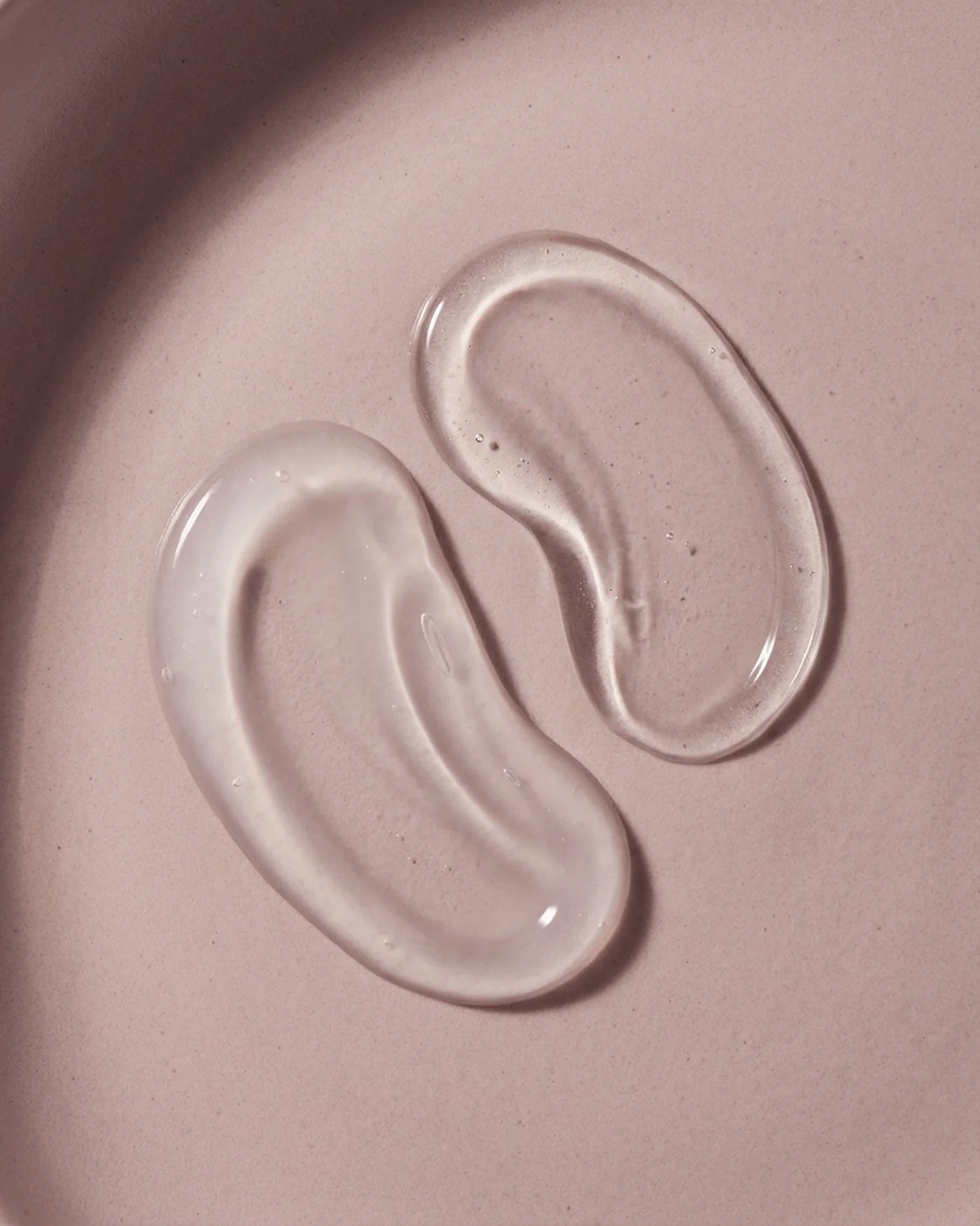 Texture macro of René Fris Anti-Frizz Shampoo and Anti-Frizz Conditioner. Two clear gel-like substances on a pink surface